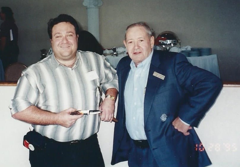 Bob Loveless in a blazer at a knife show in 1995, standing with knife maker Adam Drescher, who is holding a Loveless knife