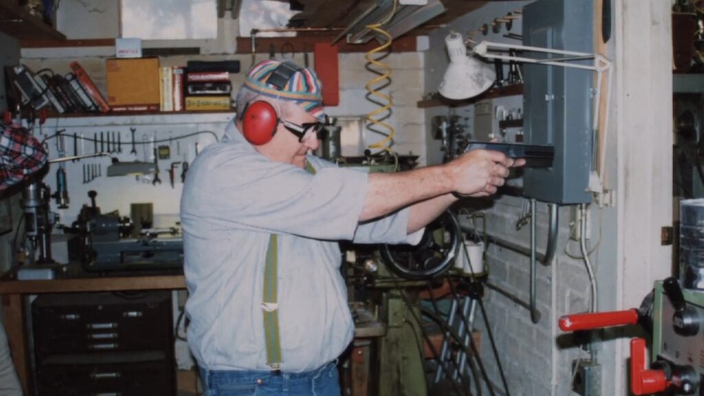 Bob Loveless in his workshop wearing ear defenders and safety glasses, aiming a pistol at a target box