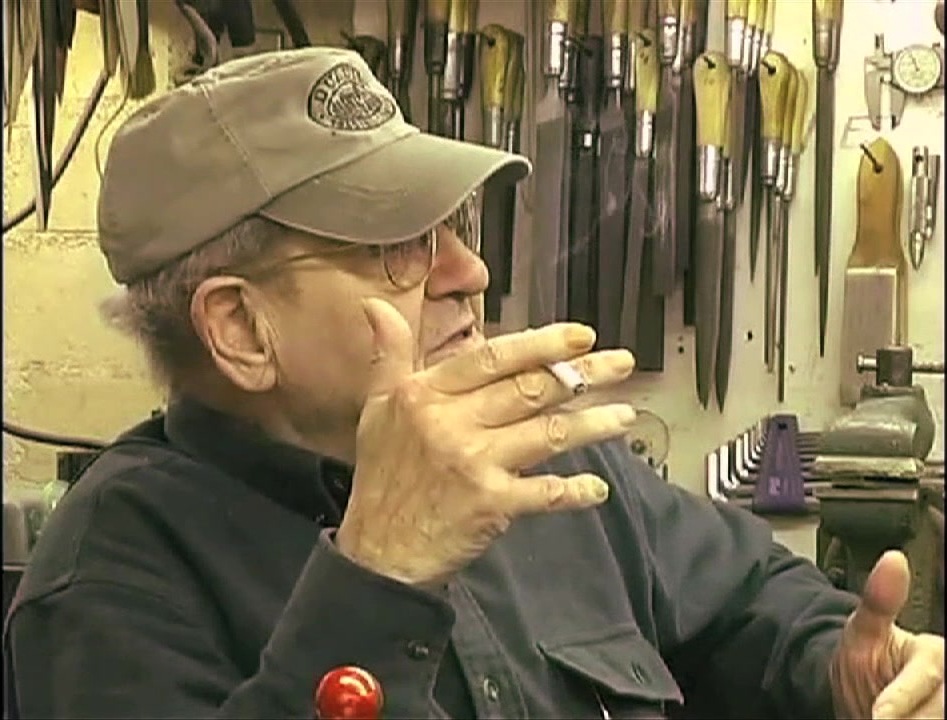 Bob Loveless in his workshop wearing a cap, with knife blade blanks and tools on the wall behind him